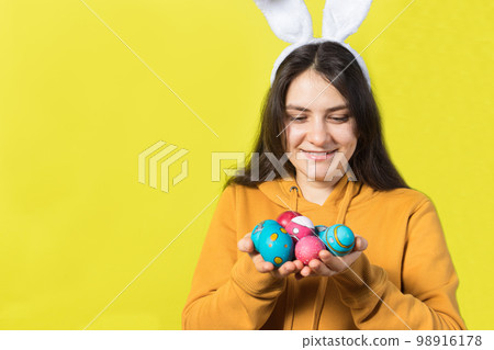 A happy brunette woman with rabbit ears on her head holds Easter eggs and smiles. Prepare for Easter, on a yellow background with copy space for text. 98916178