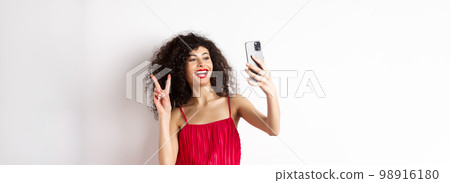 Elegant young woman in red dress taking selfie on smartphone, posing on event party, standing with mobile phone on white background 98916180