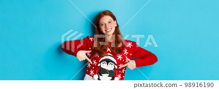 Winter holidays and Christmas Eve concept. Pretty redhead girl pointing fingers at cute xmas sweater with penguin, standing over blue background 98916190