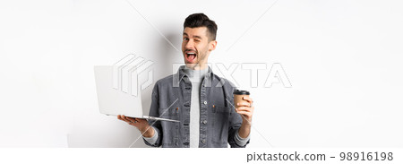 Happy male worker drinking coffee and work with laptop, winking at camera, standing on white background 98916198