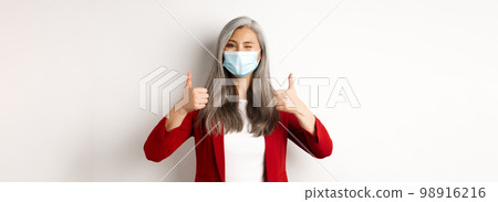 Coronavirus and business concept. Asian female entrepreneur in face mask looking cheerful, showing thumb-up in approval, white background Coronavirus and business concept. Asian female entrepreneur in face mask looking cheerful, showing thumb-up in approval, white background 98916216