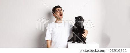 Amazed young man in glasses hugging his dog, pet owner and pug staring at upper left corner promo offer, standing over white background 98916231