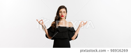Calm and patient young brunette female meditating, standing in black dress with hands in zen gesture, standing against white background 98916269