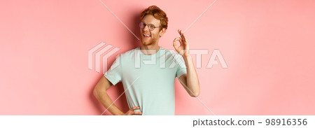 Cheerful guy with red hair and beard, wearing glasses, showing OK sign in approval and winking, smiling satisfied, standing over pink background 98916356
