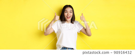 Pretty young asian woman in white t-shirt, showing thumbs up and smiling, praise good offer, recommend product, standing satisfied over yellow background 98916432