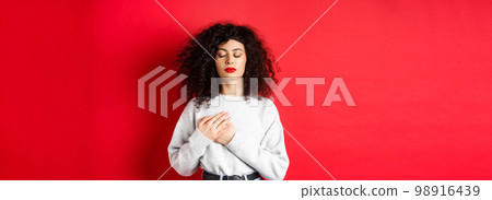 Image of calm young woman with curly hairsty, close eyes and holding hands on heart, keeping warm memories, feeling nostalgic, standing on red background 98916439