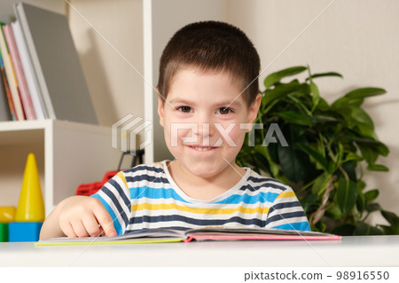 A beautiful happy child of 4 years old learns to read, sits in front of a book, looks into the camera and smiles 98916550