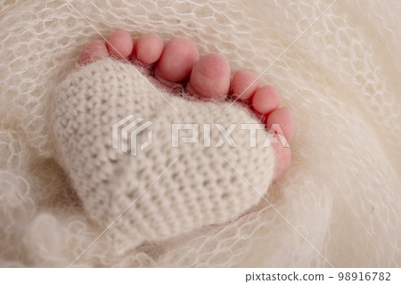 The tiny foot of a newborn baby. Soft feet of a new born in a wool white blanket. Closeup of toes, heels and feet of a newborn. Knitted heart in the legs of baby. Macro studio photography. The tiny foot of a newborn baby. Soft feet of a new born in a wool white blanket. Closeup of toes, heels and feet of a newborn. Knitted heart in the legs of baby. Macro studio photography. 98916782
