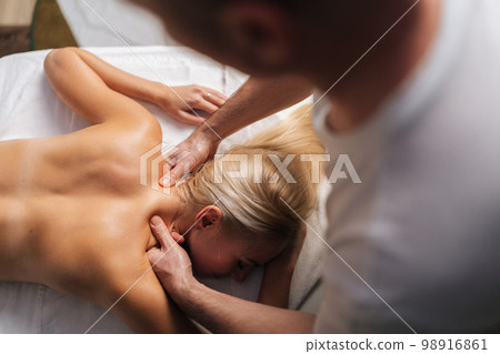 High-angle view of unrecognizable male masseur doing back and neck massage to naked female client in spa salon lying on massage table. Concept of body care and rehabilitation of spiritual peace. High-angle view of unrecognizable male masseur doing back and neck massage to naked female client in spa salon lying on massage table. Concept of body care and rehabilitation of spiritual peace. 98916861