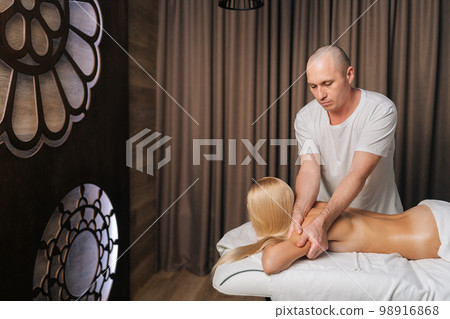 Side view of male masseur doing back and shoulder massage to blonde female client with beautiful skin lying in spa salon. Concept of body care and rehabilitation of spiritual peace. 98916868