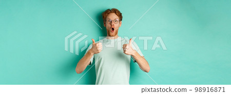 Amazed young man with red hair and beard, wearing glasses with t-shirt, showing thumbs-up and gasping in awe, checking out awesome promo offer, turquoise background Amazed young man with red hair and beard, wearing glasses with t-shirt, showing thumbs-up and gasping in awe, checking out awesome promo offer, turquoise background 98916871