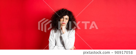 Grumpy young woman with curly hair, looking annoyed or bored at empty space, standing pensive and sad on red background Grumpy young woman with curly hair, looking annoyed or bored at empty space, standing pensive and sad on red background 98917058