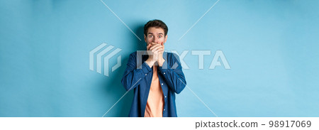 Shocked man covering mouth and gasping ambushed, witness something scary, standing on blue background Shocked man covering mouth and gasping ambushed, witness something scary, standing on blue background 98917069