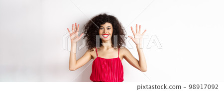 Beautiful female model with curly hair, wearing red dress, showing number ten with fingers, standing against white background 98917092