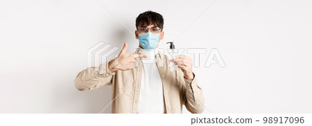 Health, covid and quarantine concept. Cheerful young guy in face mask an glasses pointing finger at bottle of antiseptic, showing good hand sanitizer, white background 98917096