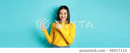Shopping concept. Attractive asian girl in trendy sweater showing advertisement, pointing fingers right and smiling, recommend promotion deal, blue background 98917110