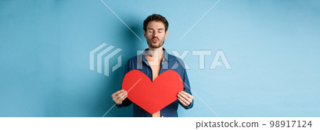 Romantic boyfriend with valentines red heart close eyes, pucker lips and waiting for kiss on lovers day, standing against blue background Romantic boyfriend with valentines red heart close eyes, pucker lips and waiting for kiss on lovers day, standing against blue background 98917124