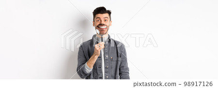 Handsome positive guy showing white perfect smile with magnifying glass, standing against white background 98917126