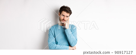 Worried young man with beard in sweatshirt, looking away pensive and thinking, standing troubled against white background Worried young man with beard in sweatshirt, looking away pensive and thinking, standing troubled against white background 98917127