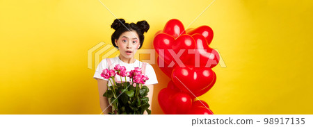Happy Valentines day. Surprised asian girl receive bouquet of pink roses from lover, looking with awe at camera, standing near red heart balloons, yellow background 98917135