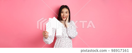 Real estate. Adult asian woman searching for home, holding house model and smiling, promo of broker company, standing over pink background Real estate. Adult asian woman searching for home, holding house model and smiling, promo of broker company, standing over pink background 98917161
