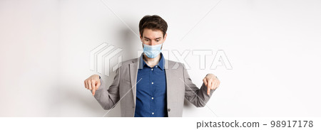 Covid-19, pandemic and business concept. Surprised guy in medical mask and suit looking and pointing down with popped eyes, gasping amazed, white background Covid-19, pandemic and business concept. Surprised guy in medical mask and suit looking and pointing down with popped eyes, gasping amazed, white background 98917178