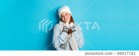 Winter and holidays concept. Cute redhead girl blushing and looking away dreamy, wearing warm beanie, sweater and scarf with gloves, standing over blue background Winter and holidays concept. Cute redhead girl blushing and looking away dreamy, wearing warm beanie, sweater and scarf with gloves, standing over blue background 98917181