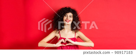 Lovers day. Beautiful woman celebrating valentines, showing heart sign and smiling, standing in romantic red dress on studio background Lovers day. Beautiful woman celebrating valentines, showing heart sign and smiling, standing in romantic red dress on studio background 98917183