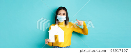 Covid-19, pandemic and real estate concept. Cheerful asian woman smiling in medical mask, showing paper house cutout, recommend agency for buying property, blue background Covid-19, pandemic and real estate concept. Cheerful asian woman smiling in medical mask, showing paper house cutout, recommend agency for buying property, blue background 98917197