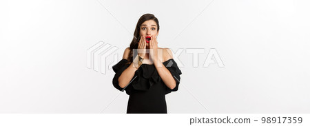 Fashion and beauty. Image of attractive female model in black dress reacting to announcement, looking amazed at camera, standing surprised over white background Fashion and beauty. Image of attractive female model in black dress reacting to announcement, looking amazed at camera, standing surprised over white background 98917359