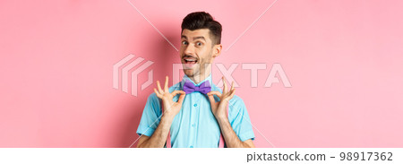 Classy handsome man fixing bow-tie on neck and smiling, getting ready for date or party, standing on pink background Classy handsome man fixing bow-tie on neck and smiling, getting ready for date or party, standing on pink background 98917362