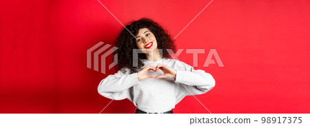 Beautiful young woman with curly hair showing heart gesture, say I love you and smile romantic at camera, standing on red background 98917375