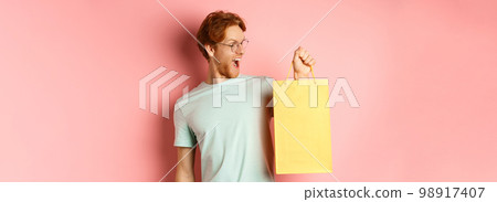 Handsome young man buying presents, holding shopping bag and looking amused, standing over pink background 98917407