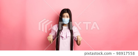 Healthy people and covid-19 pandemic concept. Sad asian woman pointing fingers down, frowning upset, standing against pink background 98917558