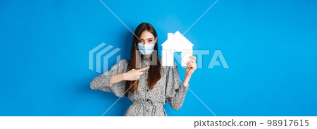 Real estate, covid-19 and pandemic concept. Smiling female model in medical mask pointing at paper house cutout, standing on blue background Real estate, covid-19 and pandemic concept. Smiling female model in medical mask pointing at paper house cutout, standing on blue background 98917615