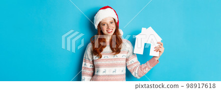 Holiday promos and real estate concept. Cheerful redhead woman in santa hat holding paper house in hand and smiling, standing in sweater against blue background Holiday promos and real estate concept. Cheerful redhead woman in santa hat holding paper house in hand and smiling, standing in sweater against blue background 98917618