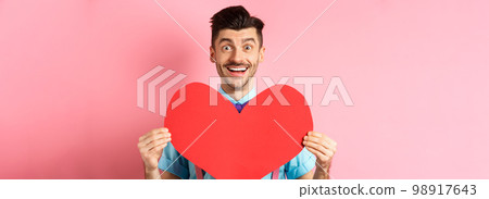 Valentines day concept. Romantic man falling in love, showing big red heart cutout and smiling, standing on pink background Valentines day concept. Romantic man falling in love, showing big red heart cutout and smiling, standing on pink background 98917643
