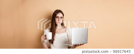 Tired working girl looking at laptop bored, drinking coffee while using computer for work, standing on beige background Tired working girl looking at laptop bored, drinking coffee while using computer for work, standing on beige background 98917645