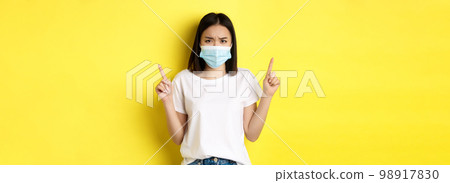 Covid-19, pandemic and social distancing concept. Disappointed asian girl in medical mask, frowning upset and pointing fingers up at logo, standing over yellow background Covid-19, pandemic and social distancing concept. Disappointed asian girl in medical mask, frowning upset and pointing fingers up at logo, standing over yellow background 98917830