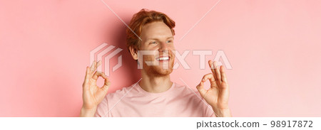 Close up of handsome redhead man in t-shirt, smiling satisfied and looking left, showing okay signs in approval, praising awesome product, standing over pink background Close up of handsome redhead man in t-shirt, smiling satisfied and looking left, showing okay signs in approval, praising awesome product, standing over pink background 98917872