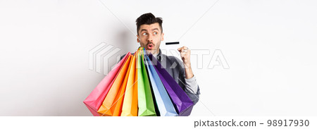 Excited young man buying in shop with plastic credit card, holding colorful shopping bags and looking aside at logo, standing against white background 98917930