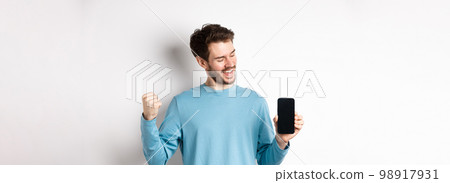 E-commerce and shopping concept. Lucky man winning online, showing empty smartphone screen and triumphing, saying yes with fist pump, standing over white background E-commerce and shopping concept. Lucky man winning online, showing empty smartphone screen and triumphing, saying yes with fist pump, standing over white background 98917931