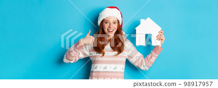 Holiday promos and real estate concept. Satisfied female model with red wavy hair, wearing santa hat and sweater, showing paper house model and thumbs-up, blue background Holiday promos and real estate concept. Satisfied female model with red wavy hair, wearing santa hat and sweater, showing paper house model and thumbs-up, blue background 98917957