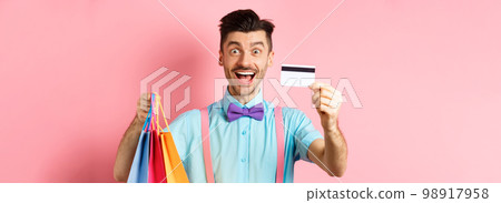 Happy young man giving his credit card and holding shopping bags, buying with discounts, standing on pink background and smiling Happy young man giving his credit card and holding shopping bags, buying with discounts, standing on pink background and smiling 98917958