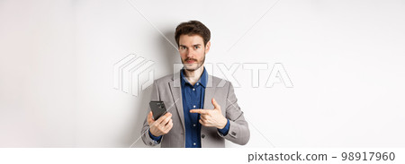 Caucasian business man in suit pointing at smartphone, showing on mobile phone, white background Caucasian business man in suit pointing at smartphone, showing on mobile phone, white background 98917960