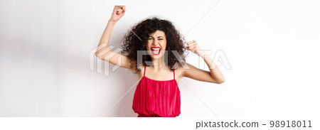 Cheerful emotive woman with curly hair, red dress, raising hands up and chanting, rooting for team, shouting wanting to win, standing on white background Cheerful emotive woman with curly hair, red dress, raising hands up and chanting, rooting for team, shouting wanting to win, standing on white background 98918011