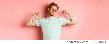 Confident young man with red hair, wearing summer sunglasses and t-shirt, showing strong and fit body muscles, flex biceps and staring cool at camera, pink background Confident young man with red hair, wearing summer sunglasses and t-shirt, showing strong and fit body muscles, flex biceps and staring cool at camera, pink background 98918021