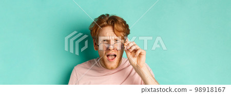 Close up portrait of redhead guy take-off glasses and looking confused at something strange, standing shocked over turquoise background 98918167