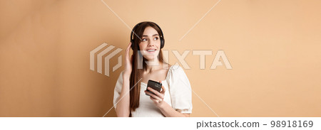 Attractive girl listening music in wireless headphones, holding mobile phone and looking aside with dreamy smile, beige background 98918169