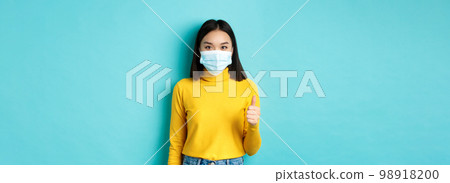 Covid-19, social distancing and pandemic concept. Young woman in medical mask showing thumbs up in approval, saying yes, standing over blue background Covid-19, social distancing and pandemic concept. Young woman in medical mask showing thumbs up in approval, saying yes, standing over blue background 98918200